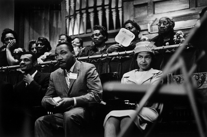 Black-and-white photo of Martin Luther King Jr. and Rosa Parks seated side by side at a civil rights gathering, with supporters seated behind them.