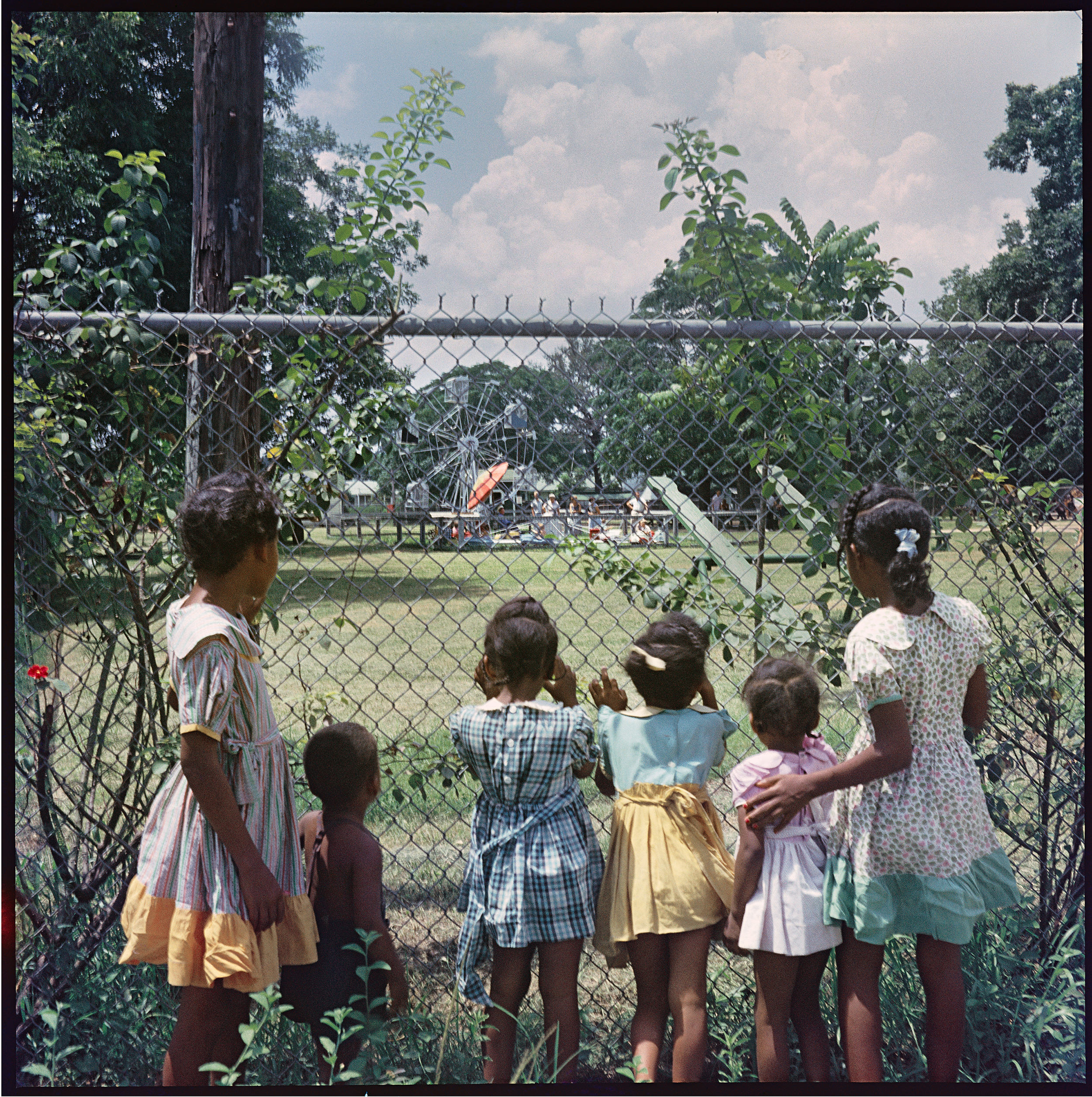 Black children stand behind a fence looking into a park playground.  Photo taken in Alabama by: Gordon Parks